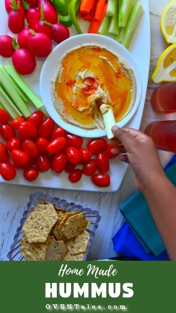 A white platter with hummus surrounded by colorful veggies. A celery stick is being dipped in the hummus.