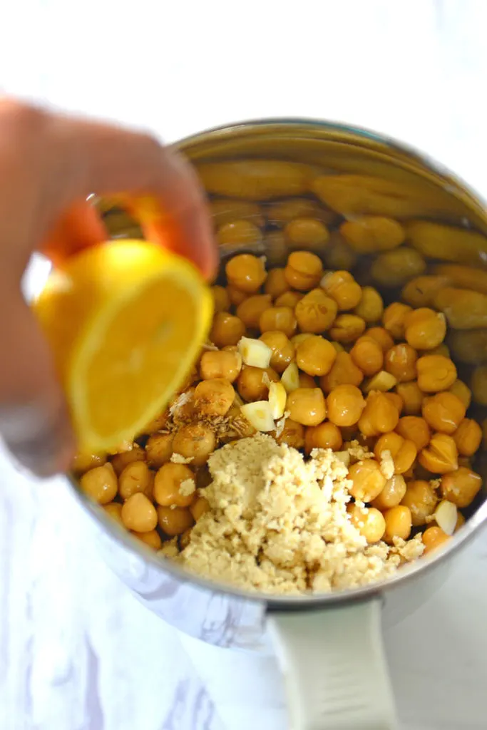 Chickpeas, tahini, pinch of cumin,    and garlic in a blender bowl  with lemon being squeezed over it.