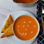 A bowl of creamy Tomato Soup with a 2 triangular slices of toast and a pot of soup next to it