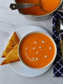 A bowl of creamy Tomato Soup with a 2 triangular slices of toast and a pot of soup next to it