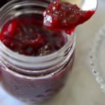 A spoon scooping red jelly from a bottle a bottle with image caption - Plum Chutney.