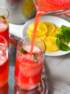 A jug of Watermelon Agua Fresca pouring into a tall glass with garnishes of lemon and mint in teh background