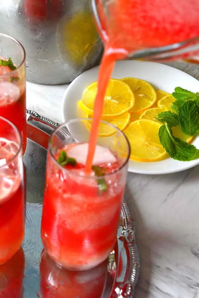 A jug of  Watermelon Agua Fresca pouring into a tall glass with garnishes of lemon and mint in teh background