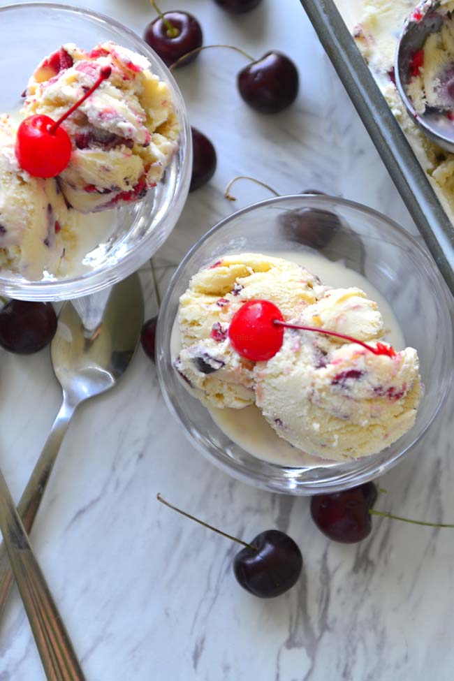 Two bowls of  cherry topped ice cream  with a few fresh cherries scattered around tehm.