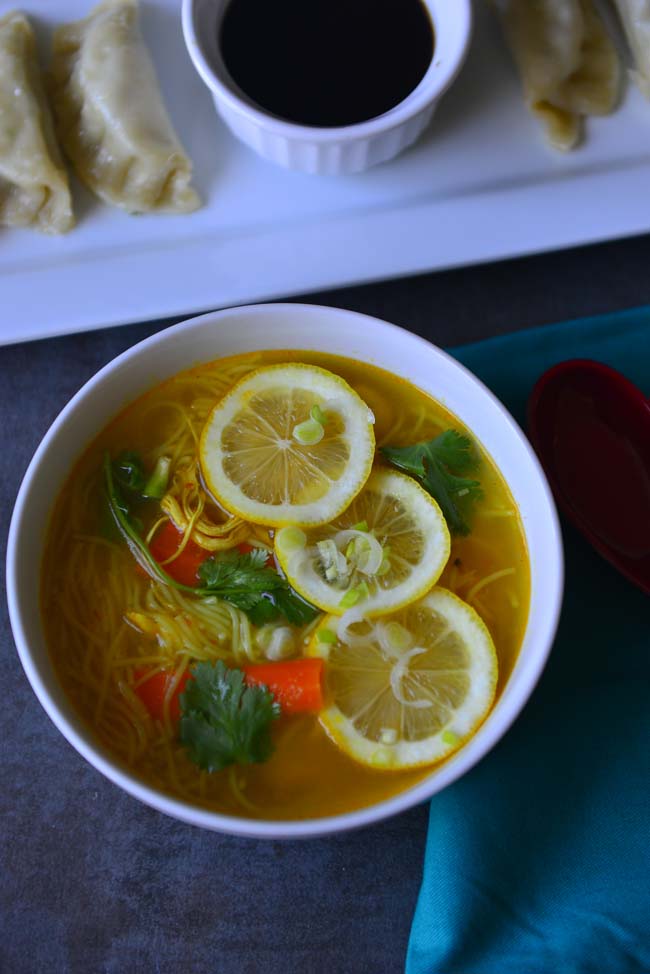 A bowl of Soup topped with lemon  and  along with a few momos and dipping sauce next to it 