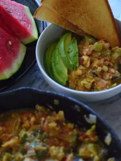 Bowl with menemen, avoccado and toast next to sliced watermelon and pan of egg scramble