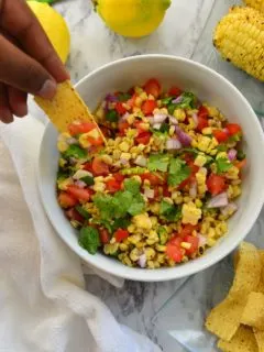 Someone trying to scoop grilled corn salsa with a chip from a white bowl