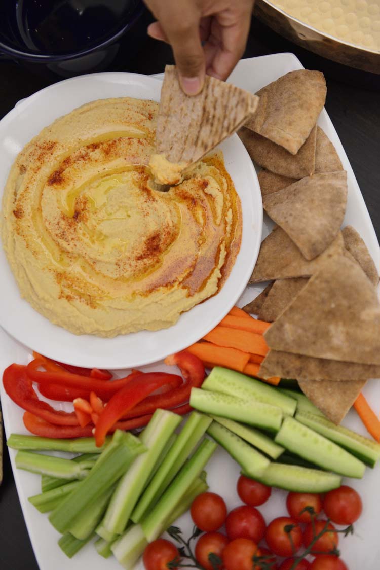 A plate of hummus  with slices of pita bread and vegetables around if.