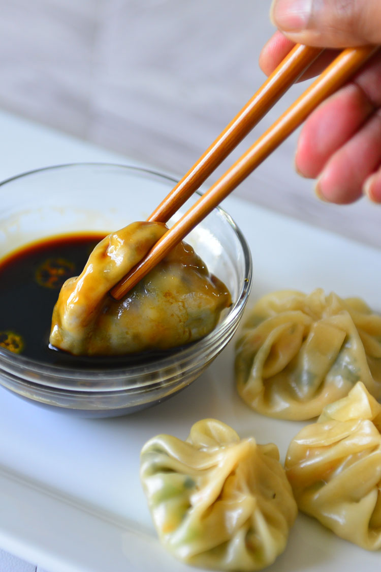 A  plate with momos and sauce set on it. One momo is being dipped in the sauce using chopsticks 