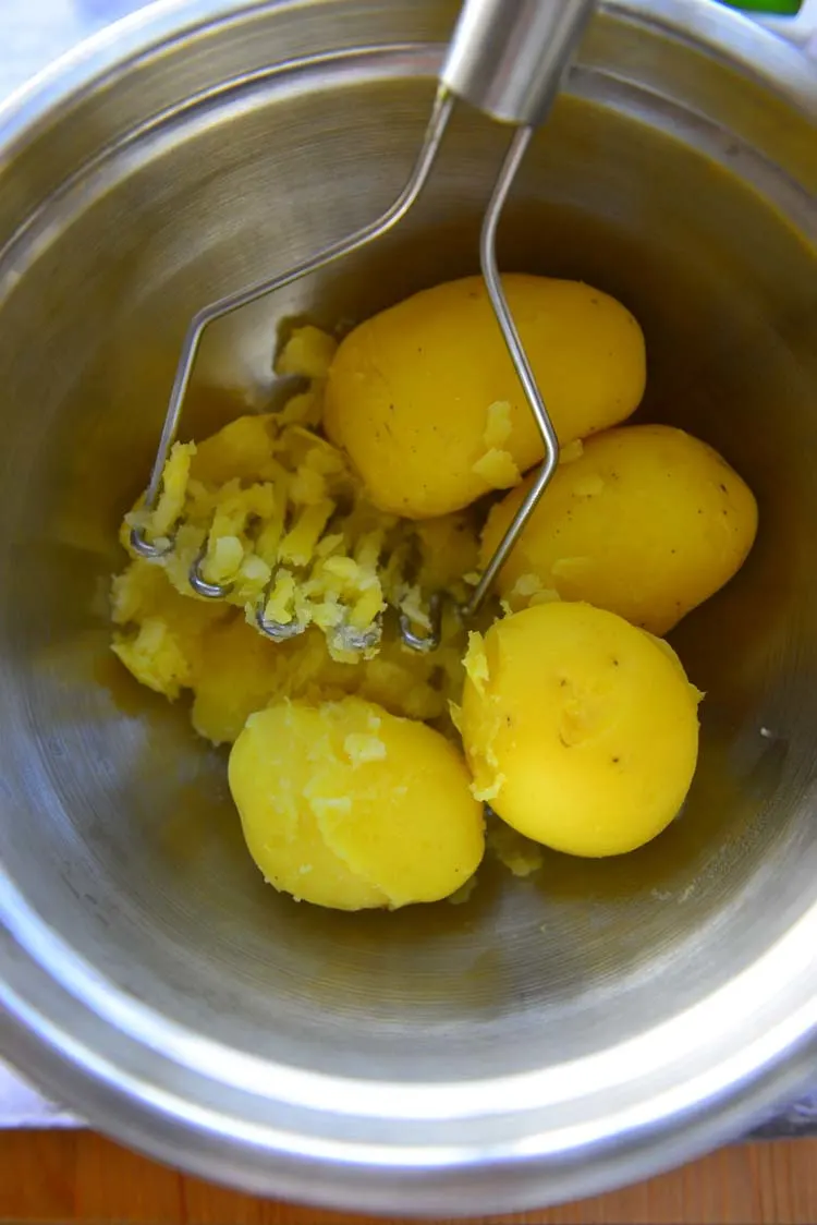 Boiled potatoes being mashed with potato masher in a steel bowl