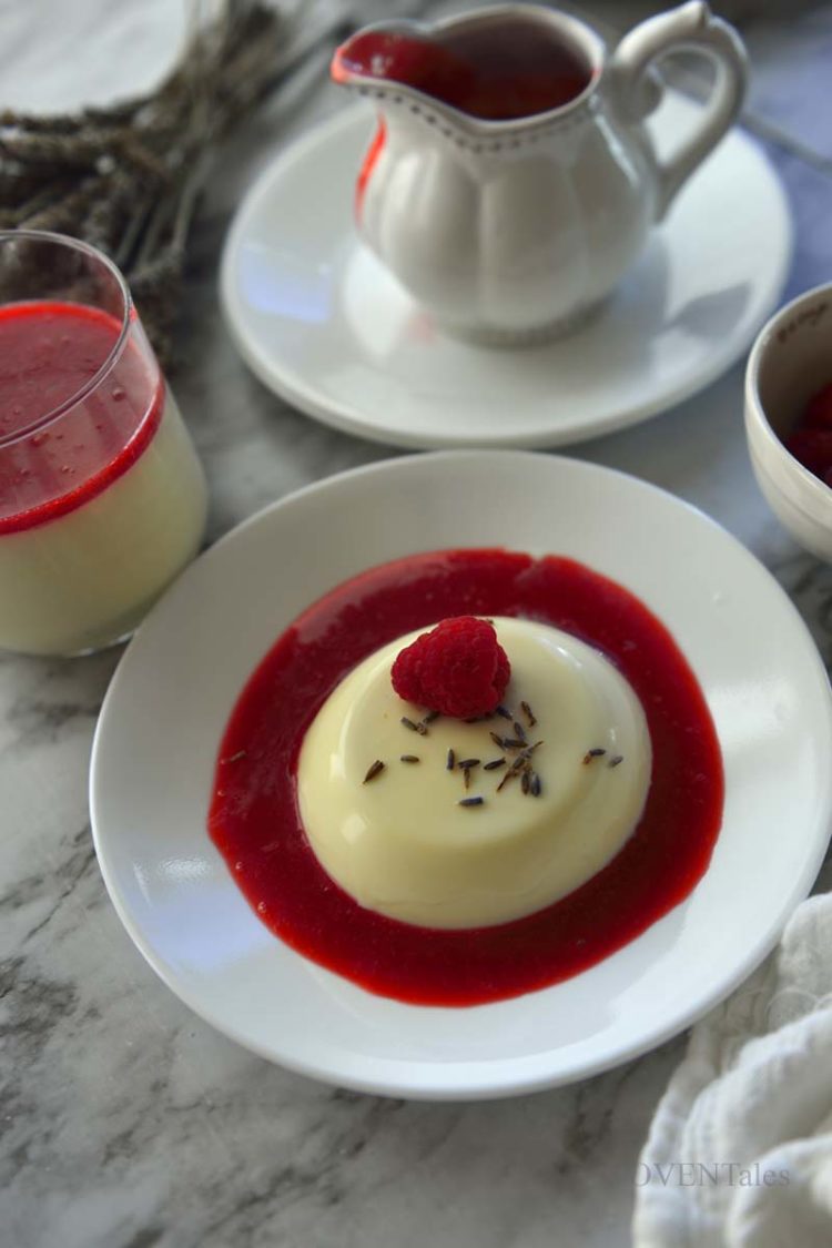 Lavender Panna Cotta in raspberry sauce on a white plate. Sauce cup and more pudding cups next to it.
