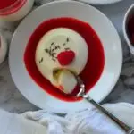 Pinnable image of lavender panna cotta on a white plate with red sauce around it . A spoon dipping into it and captioned - Lavender Panna Cotta With Raspberry Sauce