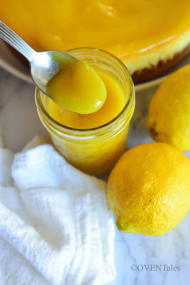 Scooping lemon curd from a bottle to show the texture, and lemons and tart next to it. 