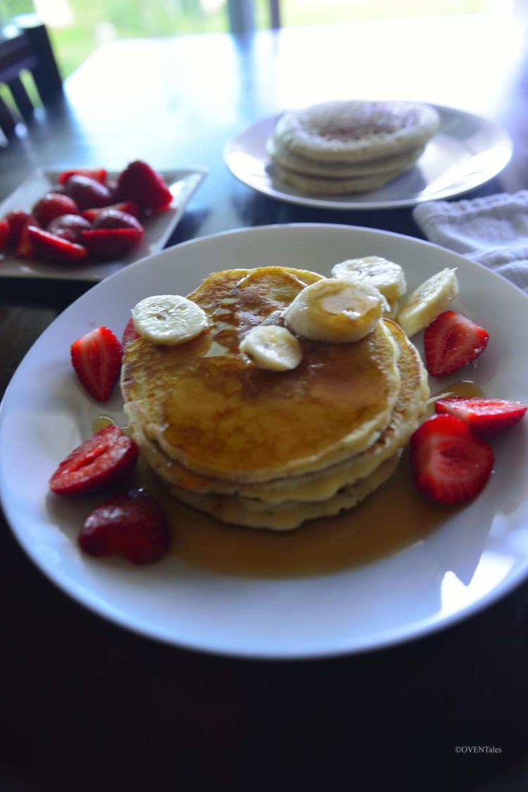 Stack of pancakes on a plate with fruits and syrup.